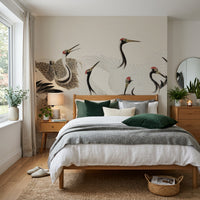 A styled bedroom interior featuring a large wall mural with black and gray cranes on a beige background, behind a wooden bed with white and gray bedding and green pillows.