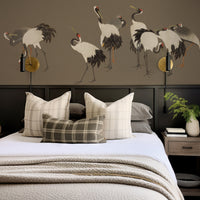 A styled bedroom scene featuring a bed with cream bedding, plaid accent pillows, and a large wall mural of cranes on a taupe wall with gold wall sconces.