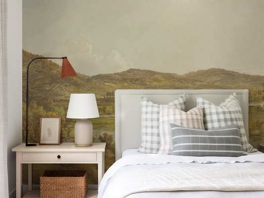 Bedroom with a bed featuring checkered and striped pillows, a lamp on a nightstand, and a scenic wall mural.