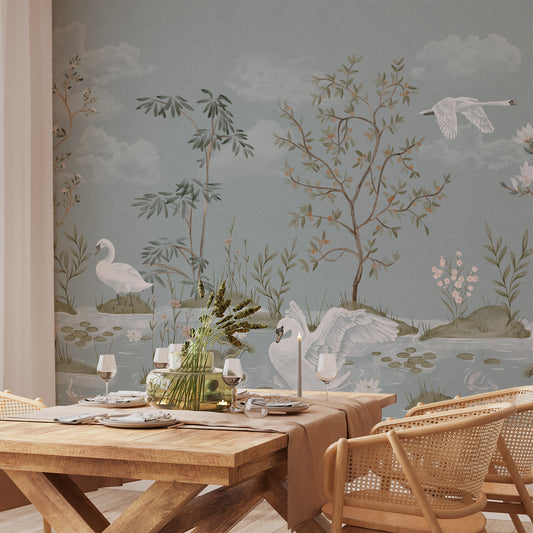 A styled dining room featuring a large wall mural with a delicate nature scene of swans, trees, and birds on a muted blue-gray background. In the foreground, a wooden dining table is set with plates, candles, and a green floral centerpiece, surrounded by woven rattan chairs.