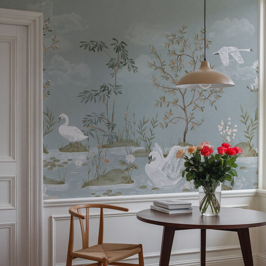 A styled interior room featuring a large wall mural with a delicate swan and landscape motif in muted blue-gray, beige, and green tones. A wooden chair, round dark wood table, vase of red flowers, and pendant light are arranged in front of the wall.