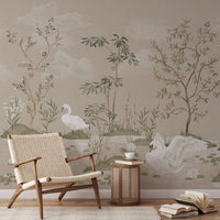 A styled interior scene featuring a large wall mural with a soft botanical landscape, including white swans, trees, and clouds on a warm beige background. A woven armchair, a small round side table with a cup and book, and scattered books are placed in front of the wall.