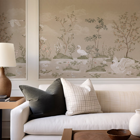 A styled interior scene showing a white sofa with two decorative pillows in front of a large wall mural or wallpaper featuring a soft landscape with swans, trees, and water in muted beige, gray, and green tones.