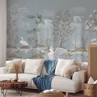 A styled living room featuring a large wall mural with a delicate landscape design of swans, trees, and clouds in muted blue-gray tones. In front of the mural is a cream-colored sofa with neutral and blue throw pillows and a blue throw blanket, a small wooden coffee table, and a woven basket on the floor.