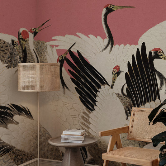 A styled interior corner featuring a large wall mural with black cranes on a dusty pink background, alongside a floor lamp, small side table with books, and a wooden chair.