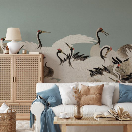 A styled living room featuring a large wall mural with elegant white cranes in black and red details against a muted sage green background. In front of the wall is a white sofa with blue and beige pillows, a light wood cabinet with a white lamp, and a low wooden coffee table with books and a vase.