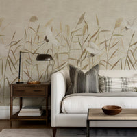A styled living room corner featuring a large-scale botanical wallpaper mural with white cranes and beige reeds on a light taupe background. The wall is decorated with a white sofa, gray and plaid throw pillows, a dark wood side table with a black lamp, and a light wood coffee table with a wooden bowl.