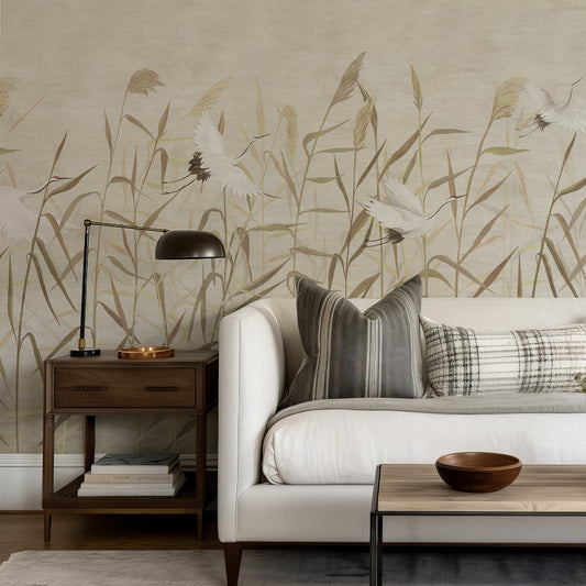 A styled living room corner featuring a large-scale botanical wallpaper mural with white cranes and beige reeds on a light taupe background. The wall is decorated with a white sofa, gray and plaid throw pillows, a dark wood side table with a black lamp, and a light wood coffee table with a wooden bowl.