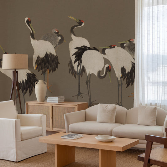 A styled living room featuring a large wall mural of elegant cranes in black, white, and gray tones against a warm taupe wall. The room is furnished with a cream-colored sofa, matching armchair, a light wood coffee table, and a wooden side cabinet with a ceramic vase.