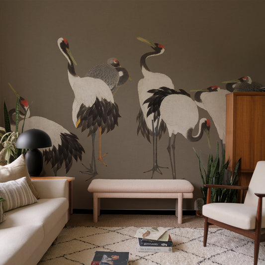 A large wall mural featuring painted cranes on a taupe wall in a styled living room with a sofa, bench, chair, rug, and plants.