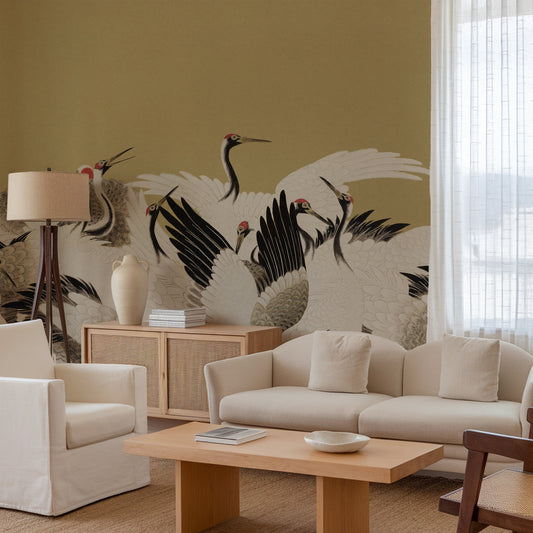 A styled living room featuring a large wall mural of black and white cranes with red beaks and feet on a warm mustard-yellow background. The room includes a cream-colored sofa, a matching armchair, a light wood coffee table, a floor lamp, and a window with sheer white curtains.