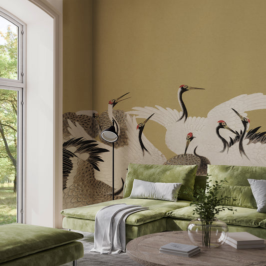 A styled living room featuring a large wall mural with elegant white cranes and brown birds on a warm beige background. The room is furnished with a green sectional sofa, a matching green ottoman, a black floor lamp, and a round wooden coffee table with books and a glass vase.