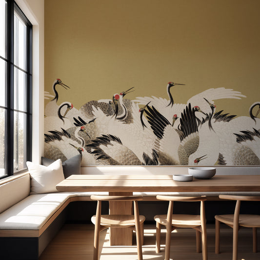 A styled interior corner with a mustard-yellow wall featuring a large mural of cranes, paired with a wooden dining table, light wood chairs, and a built-in bench with white cushions near a large window.