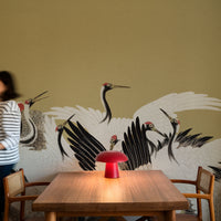 A dining area with a wooden table and chairs in front of a large wall mural depicting white cranes with black and red details. A red mushroom-shaped table lamp sits on the table, and a person in a striped shirt is partially visible on the left.