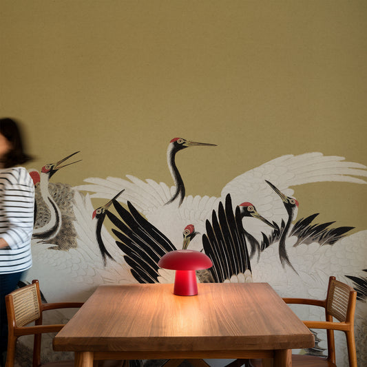 A dining area with a wooden table and chairs in front of a large wall mural depicting white cranes with black and red details. A red mushroom-shaped table lamp sits on the table, and a person in a striped shirt is partially visible on the left.