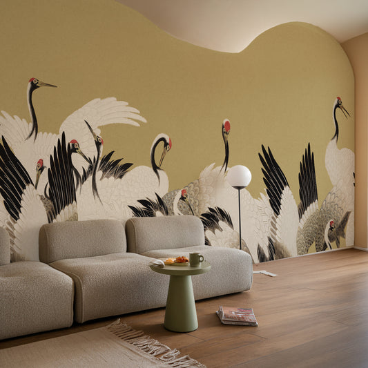 A modern living room featuring a large, curved wall mural with a traditional Chinese style illustration of white cranes with black wings and red heads on a warm beige background. In front of the wall is a light beige textured sofa, a small green side table with a mug, and a wooden floor.