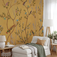 A styled interior room featuring a mustard yellow wallpaper with a botanical branch pattern and colorful birds, shown behind a white lounge chair, ottoman, side table, lamp, and floor lamp.