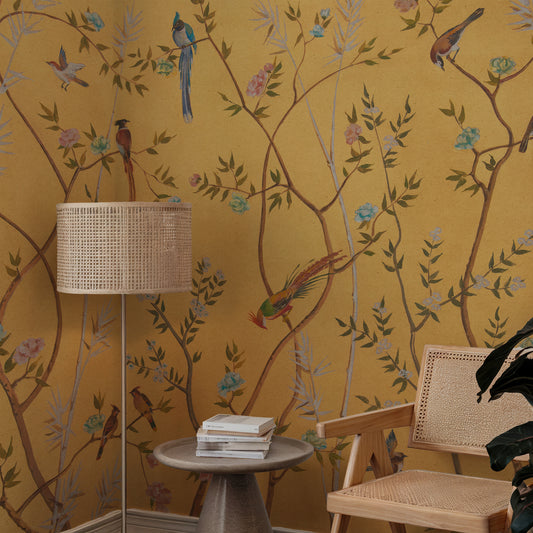 A styled interior corner featuring mustard yellow wallpaper with a botanical and bird pattern, a woven floor lamp, a small round side table with stacked books, and a light wood cane-back chair.