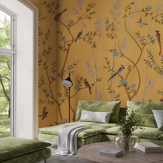 A styled living room corner featuring mustard yellow wallpaper with a delicate bird and floral branch pattern, a green velvet sectional sofa, a matching ottoman, and a round coffee table.