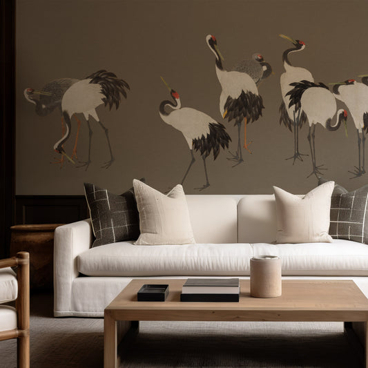 A styled living room featuring a large wall mural of elegant cranes on a taupe-brown background, above a white sofa with neutral-toned pillows and a wooden coffee table.