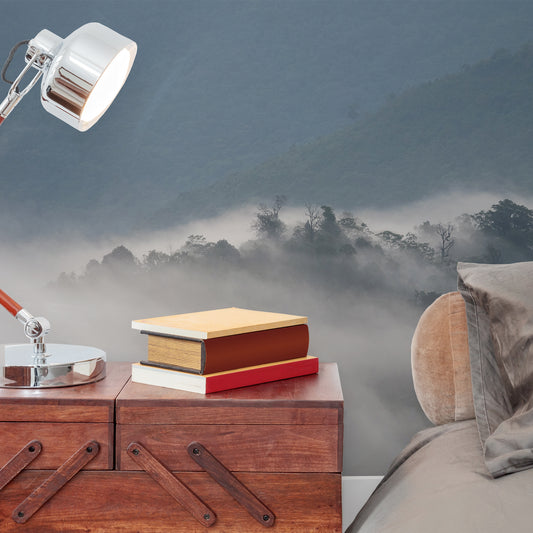 Mystical Morning haze wallpaper in bedroom with close up of grey bed and wooden side table with lamp turned on shining on a set of red books